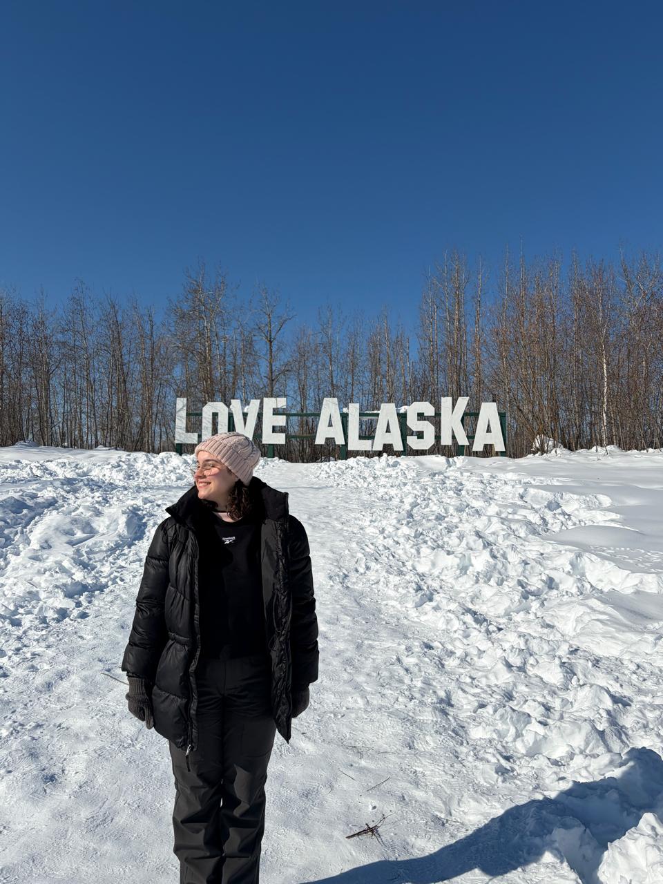 Teacher Gabi sorrindo na neve, em frente a um letreiro escrito Love Alaska